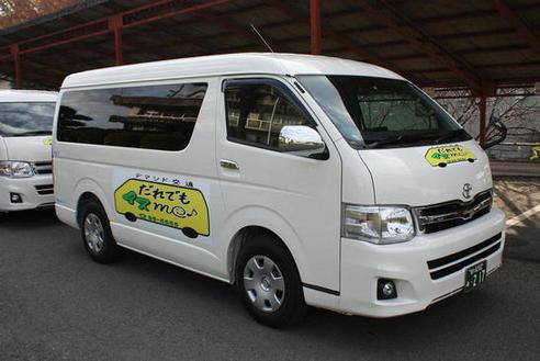 「デマンド交通」の表記の下にワゴン車の黄色い断面図がありその中に「だれでもイスme♪」とロゴが描かれている車体プリントが車体右側面と車体前面になされている中型ワゴン車が二台道路に前後に停車している写真